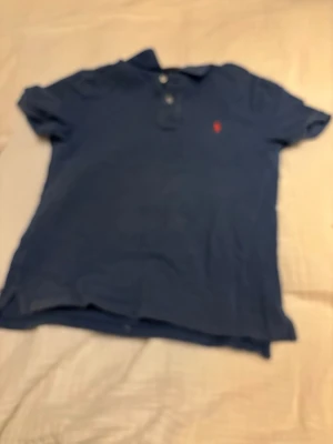 Blå pikétröja från Polo Ralph Lauren - Klassisk blå pikétröja från Polo Ralph Lauren med röd broderad logga på bröstet. Tröjan har korta ärmar, krage och knappar vid halsen. Perfekt för en avslappnad men ändå stilren look. Storlek 14 year. Hör av er vid mer info!