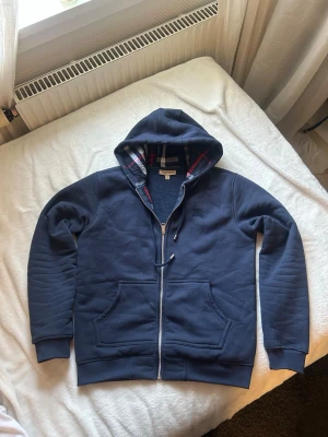 Marinblå hoodie från Burberry - Snygg marinblå hoodie från Burberry med dragkedja och klassiskt rutigt foder i huvan. Hoodien har två stora fickor fram, broderad logga på bröstet och ribbade muddar. Perfekt för dig som vill ha en stilren och exklusiv look.
