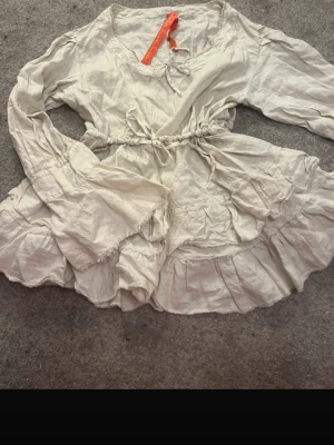 Vit volangblus från Odd Molly - Supersöt vit blus från Odd Molly med volanger och knytning i midjan. Blusen har långa ärmar med volangdetaljer och en feminin, lös passform. Tillverkad i ett lätt och luftigt material som känns skönt mot huden. Perfekt för en romantisk och bohemisk look.