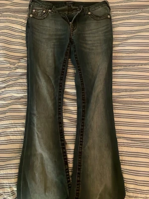 True Religion bootcut jeans blå - Säljer ett par blå bootcut jeans från True Religion med tydliga kontrastsömmar i rött och klassisk femficksdesign. Jeansen har en snygg tvättad look och ikonisk logga på bakfickan. Perfekta för dig som gillar streetwear och vill sticka ut.