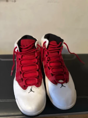 Air Jordan vita och röda sneakers - Snygga Air Jordan sneakers i vitt och rött med klassisk Jumpman-logga på tån. Skorna har högt skaft, röda snören och meshdetaljer på ovandelen. Perfekta för dig som gillar streetwear och basketinspirerad stil.