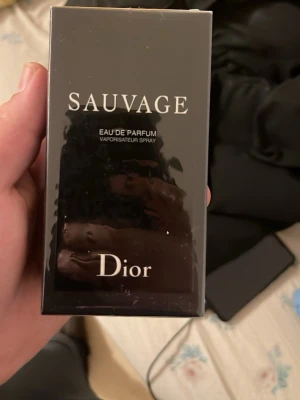Dior Sauvage Eau de Parfum - Dior Sauvage Eau de Parfum i elegant svart förpackning med vit text. Parfymen har en modern och stilren design och är känd för sin fräscha och maskulina doft. Flaskan är tillverkad av glas och kommer i originalkartong.