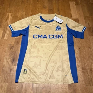 Marseille 4th kit 25/26 - Snygg Marseille matchtröja i beige med blå detaljer och tryck. Tröjan har korta ärmar, rund hals och klubbmärke på bröstet. Tillverkad i lätt och ventilerande polyester, perfekt för fotboll eller träning. Stora blå sidopaneler och sponsortryck framtill.