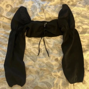 Croppad blus med puffärmar - Trendig svart croppad blus med långa puffärmar och snörning framtill. Toppen har en fyrkantig urringning och elastisk rygg för skön passform. Perfekt att styla med höga jeans eller kjol för en cool look.