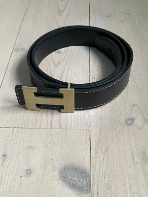 Svart H skärp med guldspänne - Snyggt svart skärp i skinn med ett stort H-format spänne i guld. Skärpet har vita kontrastsömmar längs kanterna som ger en stilren look. Perfekt accessoar för att lyfta din outfit. Knappt använt och i bra skick 110 cm