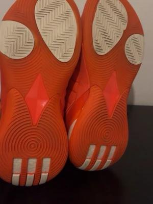 Adidas Harden orange sneakers - Snygga och sportiga sneakers från Adidas Harden-serien i en stark orange färg med vita detaljer. Skorna har en hög siluett och robust sula med greppmönster, perfekta för dig som gillar basketstil och vill sticka ut. Materialet är en mix av textil och syntet.