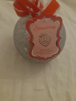 Strawberry handlotion i silverkula - Säljer en handlotion med jordgubbsdoft i en rund, glittrig silverfärgad förpackning formad som en julkula. Produkten är parabenfri och rymmer 300 ml. Dekorerad med röd rosett och etikett med jordgubbsmotiv.