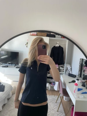 Mörkblå pikétopp från Peak Performance - Snygg mörkblå pikétopp från Peak Performance med klassisk krage och knappar framtill. Toppen har korta ärmar och en liten vit logga på bröstet. Perfekt till jeans eller kjol för en sportig och clean look.