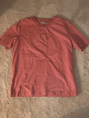 Rosa t-shirt med hjärtdetalj CLOEY - Söt rosa t-shirt från CLOEY med en unik hjärtformad detalj på . T-shirten har rund halsringning och är gjord i mjukt bomullstyg. Perfekt för dig som vill ha en enkel men ändå personlig stil.