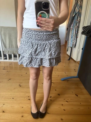 Vit kortkjol med volanger och blommor🫶🏻 - Supersöt vit kortkjol med svart blommigt mönster och dubbla volanger. Kjolen har en bekväm resår i midjan och är perfekt för varma dagar. Lätt och luftig, passar till både sneakers och sandaler. Så söt!!!!🤍🤍