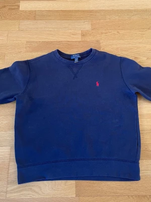 Mörkblå sweatshirt från Polo Ralph Lauren - Klassisk mörkblå sweatshirt från Polo Ralph Lauren med rund halsringning och den ikoniska röda logotypen broderad på bröstet. Tröjan har ribbade muddar vid ärmslut och nederkant, perfekt för en avslappnad och stilren look. Junior L i storlek.