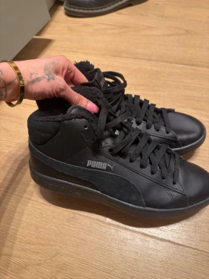 Svarta Puma sneakers med foder - Snygga svarta sneakers från Puma i mid-modell med mjukt foder. Skorna har klassisk Puma-logga på sidan, svarta skosnören och ovandel i både mocka och syntetmaterial. Perfekta för dig som vill ha stilrena och bekväma sneakers med lite extra värme.