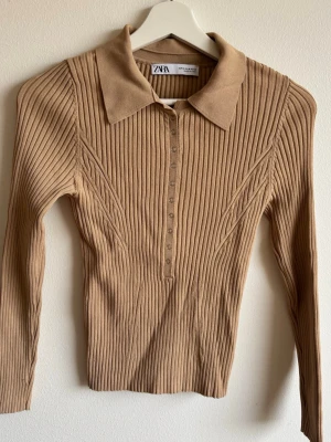 Beige ribbstickad tröja från Zara - Snygg beige ribbstickad tröja från Zara med lång ärm och klassisk krage. Tröjan har knappar framtill och är tillverkad i ett mjukt, stretchigt material som sitter tajt. Perfekt till jeans eller kjol för en clean och trendig look.