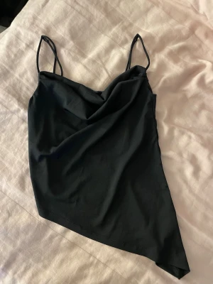 Svart asymmetrisk linnetopp - Säljer en svart linnetopp med tunna axelband och snygg asymmetrisk nederkant. Toppen har en draperad urringning och är perfekt för dig som gillar trendiga detaljer. Materialet känns mjukt och lätt mot huden.