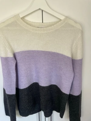 Randig stickad tröja från H&M - Säljer en stickad tröja från H&M med breda blockränder i vitt, ljuslila och mörkgrått. Tröjan har rund halsringning och långa ärmar. Perfekt för dig som gillar en avslappnad och trendig stil.