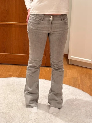 Grå bootcut jeans med dubbla knappar - Snygga grå jeans med bootcut-modell och dubbla knappar i midjan. Klassisk femficksdesign och låg midja. Jeansen är i ett mjukt och stretchigt denimtyg som ger en avslappnad men trendig look.