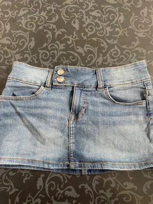 Supermini jeanskjol med dubbla knappar  - Snygg blå jeanskjol i kort modell med dubbla knappar framtill och klassiska fickor både fram och bak. Kjolen har bälteshällor och är gjord i ett slitstarkt denimtyg. Perfekt för dig som gillar en avslappnad och trendig stil.