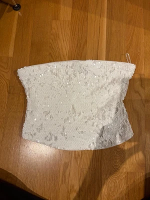 Vit paljettprydd tubtopp - Snygg vit tubtopp täckt med glittriga paljetter. Toppen är axelbandslös och har en lätt stretchig passform + gummi över så den sitter på plats. Insidan är fodrad med mjukt tyg