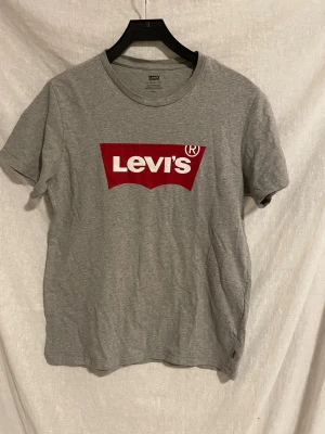 Grå Levi's t-shirt med röd logga - Snygg grå t-shirt från Levi's med klassisk röd logga på bröstet och liten röd etikett i sidan. T-shirten är i mjuk bomull och har rund hals och korta ärmar. Perfekt för en avslappnad och stilren look.