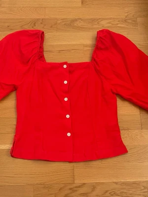 Röd blus med puffärmar och knappar - Trendig röd blus med puffiga långärmar och vita knappar framtill. Fyrkantig urringning och figurnära passform ger en modern siluett. Perfekt för dig som vill sticka ut med färg och volym. Materialet känns mjukt och lätt, troligen bomull eller bomullsblandning.
