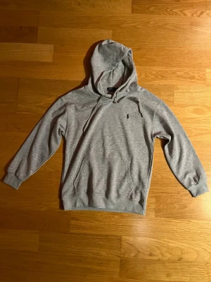 Grå hoodie från Polo Ralph Lauren - Snygg grå hoodie från Polo Ralph Lauren med klassisk huva och dragsko. Tröjan har det ikoniska broderade logotypen på bröstet och en känguruficka framtill. Tillverkad i mjuk bomullsblandning för maximal komfort och stil.