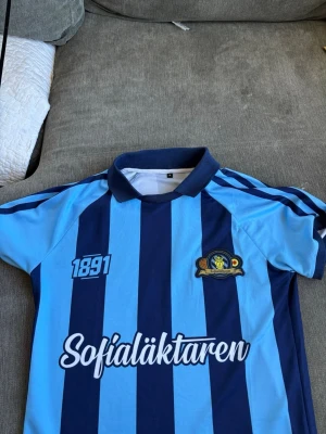 Blårandig Djurgården matchtröja - Säljer en klassisk blårandig Djurgården matchtröja med klubbmärke, årtalet 1891 och texten Sofialäktaren på framsidan. Tröjan har korta ärmar, pikékrage och texten Stockholmslaget i nacken. Perfekt för match eller supporterhäng.