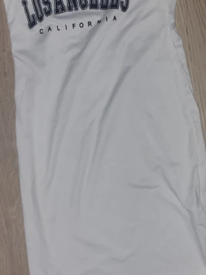 Vit kort klänning Los Angeles-tryck - Vit kort klänning med trycket 'Los Angeles California' i marinblått över bröstet. Klänningen har en enkel, rak passform och är ärmlös. Perfekt för en avslappnad och trendig look.