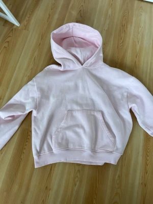 Ljusrosa hoodie med magficka - Säljer en ljusrosa hoodie i mjukt bomullsmaterial. Hoodien har en klassisk huva och en stor magficka framtill. Ribbstickade muddar vid ärmslut och nederkant ger en skön passform. Perfekt för en avslappnad och trendig look. De finns några små fläckar men syns knappt