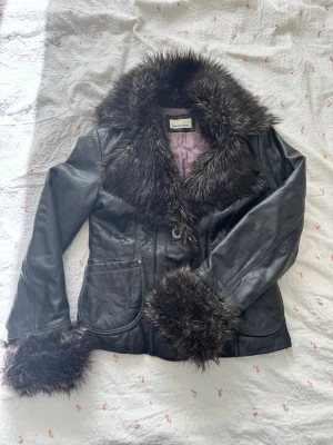 Svart skinnjacka med päls från Rock'n Blue - Cool svart skinnjacka från Rock'n Blue med stora fluffiga pälsdetaljer på krage och ärmslut. Jackan har två fickor fram och quiltat foder i lila. Perfekt för dig som vill sticka ut och ha en unik stil.