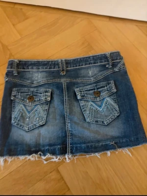 Blå jeansshorts med fransig kant - Säljer ett par blå jeansshorts med fransig kant och två bakfickor med dekorativa sömmar och knappar. Shortsen har bälteshällor och klassisk femficksdesign. Perfekta för sommardagar och enkel att matcha med olika toppar.