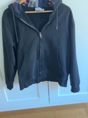 Burberry Svart  zip hoodie med rutigt foder - Svart hoodie från Burberry med dragkedja och huva. Insidan av huvan har ett rutigt foder i grått, vitt och rött. Tröjan har två fickor framtill och ribbade muddar vid ärmar och nederkant. Perfekt för att sticka ut samtidigt som en chill och avslappnad stil.