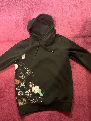 Svart hoodie med blommigt tryck - Snygg svart hoodie med huva och snörning. Unik detalj med blommigt tryck i flera färger längs ena sidan både fram och bak. Hoodien har en klassisk passform och ribbade muddar vid ärmslut och nederkant.