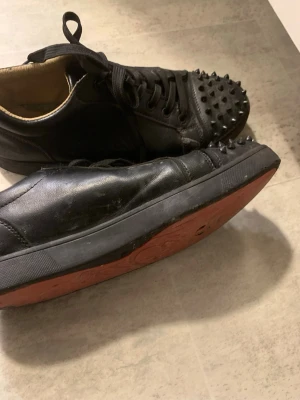 Svarta sneakers med nitar - Unika svarta sneakers i läder med coola svarta nitar på tån och klassisk röd sula. Skorna har snörning och låg profil, perfekt för dig som vill sticka ut med din stil. Insidan är beige och sulan har ett mönster för extra grepp.