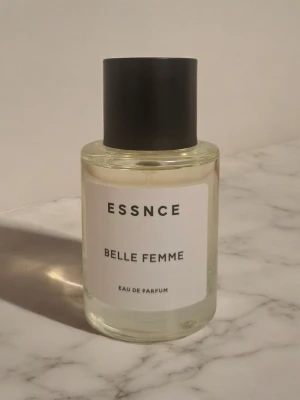 ESSNCE Belle Femme Eau de Parfum - Minimalistisk parfymflaska från ESSNCE med doften Belle Femme. Flaskan är cylindrisk i genomskinligt glas med en enkel vit etikett och svart text. Locket är svart och har en matt finish. Perfekt för dig som gillar stilrena och moderna detaljer.