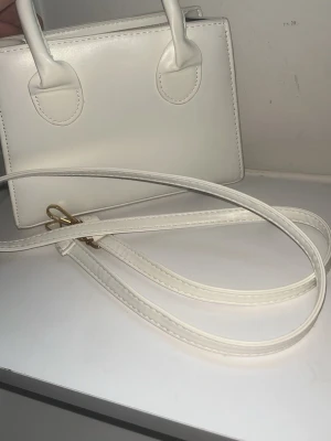 Vit minihandväska med axelrem - Stilren vit minihandväska i fyrkantig modell med både handtag och avtagbar axelrem. Väskan har guldfärgade metalldetaljer och svart innerfoder. Perfekt för dig som gillar minimalistisk design och vill ha en enkel men snygg accessoar.