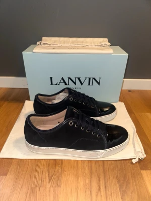 Mörkblå sneakers från Lanvin - Snygga mörkblå sneakers från Lanvin med ovandel i mocka och tåparti i blankt läder. Skorna har vita sulor, mörkblå skosnören och metallfärgade öljetter. Perfekta för dig som gillar stilrena och exklusiva sneakers. Size 43/Uk9, Box o dustbags ingår vid köp!