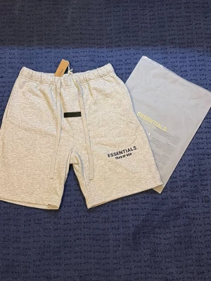 Grå Essentials shorts med snörning - Säljer ett par ljusgrå shorts från Fear of God Essentials med snörning i midjan och tryckt logga på benet. Sköna och stilrena shorts i mjukt bomullsmaterial, perfekta för sommaren. Kommer med originalpåse.