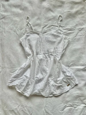 Topp  - Söt vit topp med tunna axelband och diskret prickigt mönster. Toppen har smockad överdel och en liten  logga vid fållen. Perfekt för varma dagar och enkel att styla till jeans eller kjol.