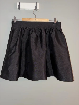 Svart ballongkjol i satin - Snygg svart ballongkjol från Gina Tricot i glansigt satinmaterial. Kjolen har en kort och voluminös form  som ger en bekväm passform. Perfekt för dig som vill ha en trendig och stilren look.