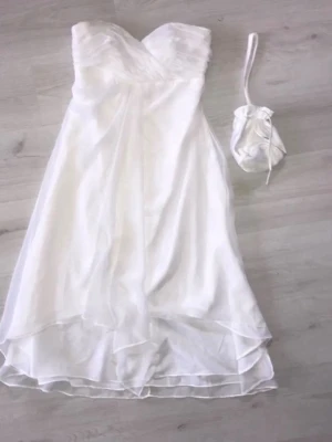 Vit axelbandslös kortklänning - Säljer en elegant vit axelbandslös kortklänning med omlott-detaljer upptill och lätt utsvängd kjol i flera lager. Klänningen har en mjuk, flowy känsla och medföljer en matchande liten väska. Perfekt för sommarens speciella tillfällen!