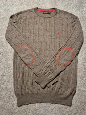 Morris stickad tröja - Säljer en brun Morris med rund halsringning och långa ärmar. Tröjan har röda detaljer på armbågarna och en liten röd logga på bröstet. Den är i bra skick förutom att den har en pytte litet håll.