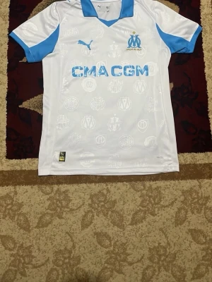 Marseille matchtröja Puma vit/blå - Säljer en vit Marseille matchtröja från Puma med ljusblå detaljer och tryck. Tröjan har klubbmärke och sponsorlogga framtill samt diskreta mönster i tyget. Perfekt för dig som älskar fotboll och vill stötta ditt lag.
