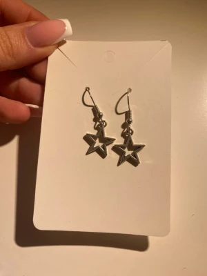 Silvriga stjärnörhängen i metall - Säljer ett par snygga örhängen i silverfärgad metall med stjärnformade hängen. Örhängena har en öppen design och hängs i örat med krok. Jag har gjort dom helt själv !!