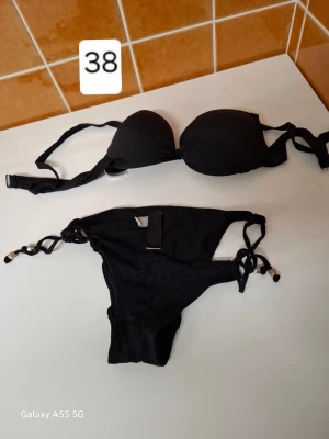 Svart bikini med snörning och detaljer - Snygg svart bikini med vadderad topp och knyt i ryggen. Underdelen har snörning i sidorna och dekorativa detaljer i metall. Perfekt för stranden eller poolen. Klassisk och stilren modell som passar till mycket.
