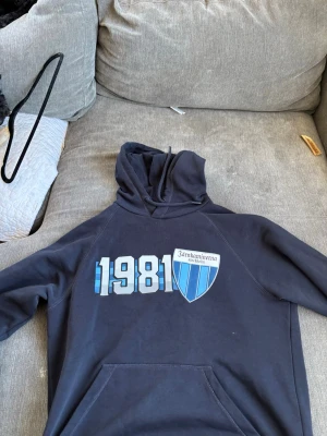 Mörkblå Särskaminerna hoodie - Mörkblå hoodie med trycket '1981' och Särskaminerna-logga i blått och vitt på bröstet. Klassisk känguruficka framtill och huva med dragsko. Perfekt för dig som vill representera laget eller bara gillar sportig stil.