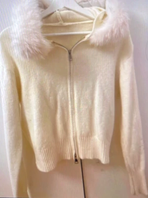 Vit hoodie med fuskpäls på huvan - Mysig vit hoodie med dragkedja och fluffig fuskpäls runt huvan. Tröjan har ribbade muddar vid ärmslut och nederkant, samt en klassisk passform. Perfekt för dig som gillar en söt och trendig stil.