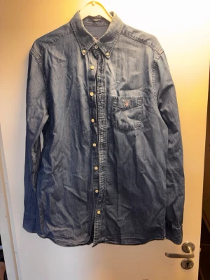 Blå jeansskjorta från Gant - Säljer en klassisk blå jeansskjorta från Gant med knappslå och bröstficka med broderad logga. Skjortan har lång ärm, button-down krage och vita knappar. Perfekt till ett avslappnat men snyggt uttryck.