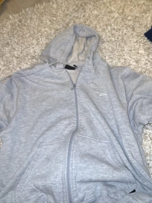 Ljusgrå hoodie från D-Way - Ljusgrå hoodie från D-Way med dragkedja framtill och två fickor. Klassisk design med huva och diskret logga på bröstet. Tillverkad i mjukt och bekvämt material som passar perfekt till en avslappnad stil.