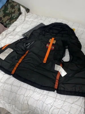 Svart dunjacka med orange detaljer - Snygg svart dunjacka från Parajumpers med orange och gula detaljer. Jackan har huva, flera innerfickor och quiltad design. Perfekt för kalla vinterdagar och har en sportig look med praktiska dragkedjor och justerbara detaljer. Problem med dragkedjan finns bild på den men de går och fixas! 