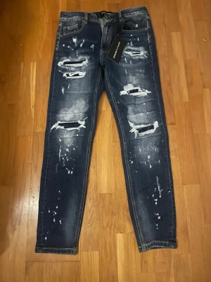 Mörkblå slitna jeans storlek M - Säljer ett par mörkblå s jeans med kraftiga slitningar och hål på framsidan samt vita färgstänk för en trendig look. Jeansen har fem fickor och normal midja. Perfekta för dig som gillar streetstyle och vill sticka ut. Storlek M